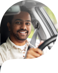 A person driving a car, wearing a light-colored shirt and a seatbelt. The background shows the car's interior and a blurred view outside.